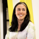 Milly Shaw: white young woman with dark hair and a white streak. She's smiling. She wears a white blouse and is front of a yellow and white background.