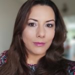 Elena Gorman is a white presenting woman with olive skin, brown eyes, and medium length dark brown hair.