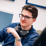 James Liddell is shown in a profile. He presents as male, has light skin, short brown hair and glasses. He is wearing a blue striped polo shirt, a silver watch, and has headphones around his neck. He's looking at something.