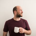 Martin, a white man with a beard, short brown hair, and bald on top. He’s looking to his left with a smile on his face and a cup of tea in one hand.