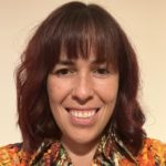 Vicky Teinaki has fair skin with freckles, shoulder-length dark red-tined hair, and dark eyes. She is smiling