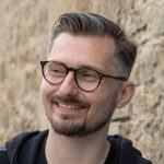 Tom Moran is a pale skinned man with short light brown styled hair and light facial hair. He is wearing glasses and smiling.