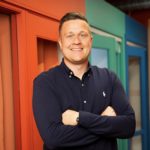 Mark Hall is a light skinned man with hair that is short, light coloured and slicked back. He is smiling, wearing a dark polo shirt and has his arms folded.