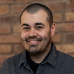 Craig Abbott smiles. He has short cropped hair, faical stubble, dark eyes and medium brown skin, and is wearing a grey shirt. He stands in front of a brick wall