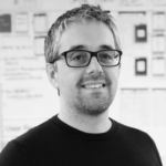 Grayscale photo of a light skinned man with light short hair, glasses, light facial stubble and a black shirt