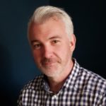 Man with light skin, grey slicked-back hair, light facial hair and a check shirt.